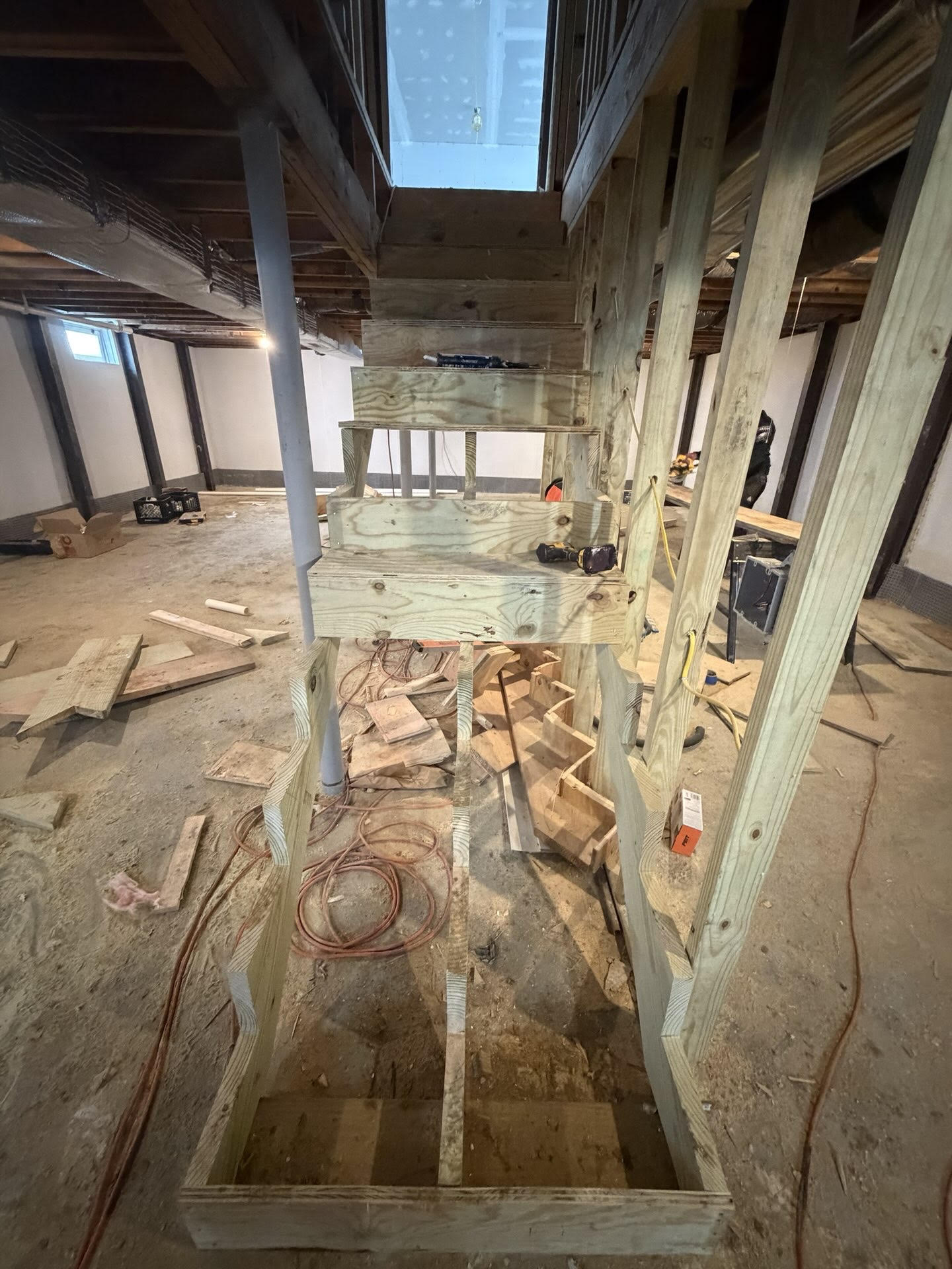 Wooden stairs under construction in a basement with exposed beams. Tools and wood planks scattered around, creating a work-in-progress atmosphere.